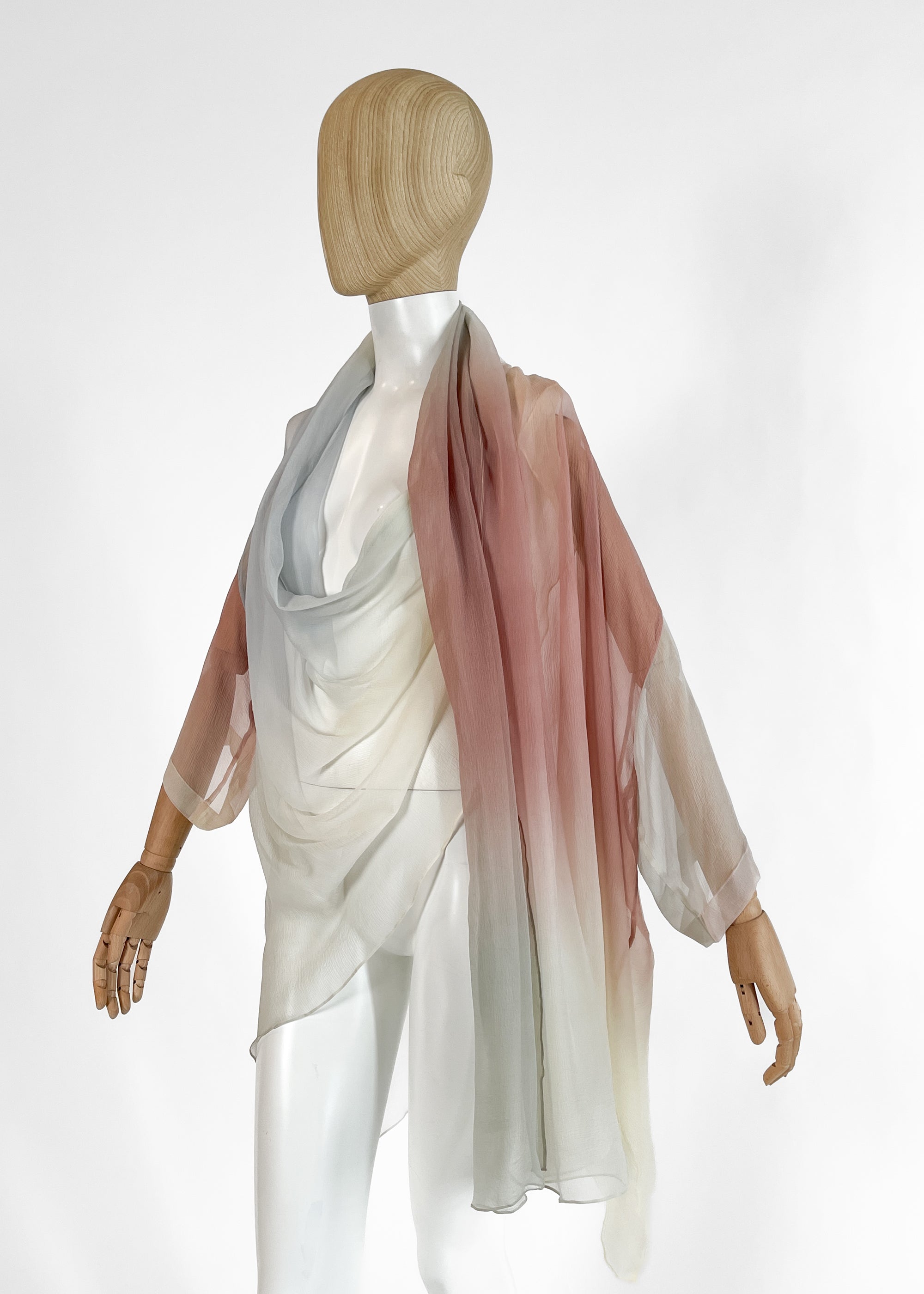 “A 1989 Romeo Gigli sheer silk kimono-style wrap top draped on a mannequin. Features a soft pastel gradient transitioning from pale blue and grey to rose pink and ivory, with a draped cowl neckline and wide, fluid sleeves.”