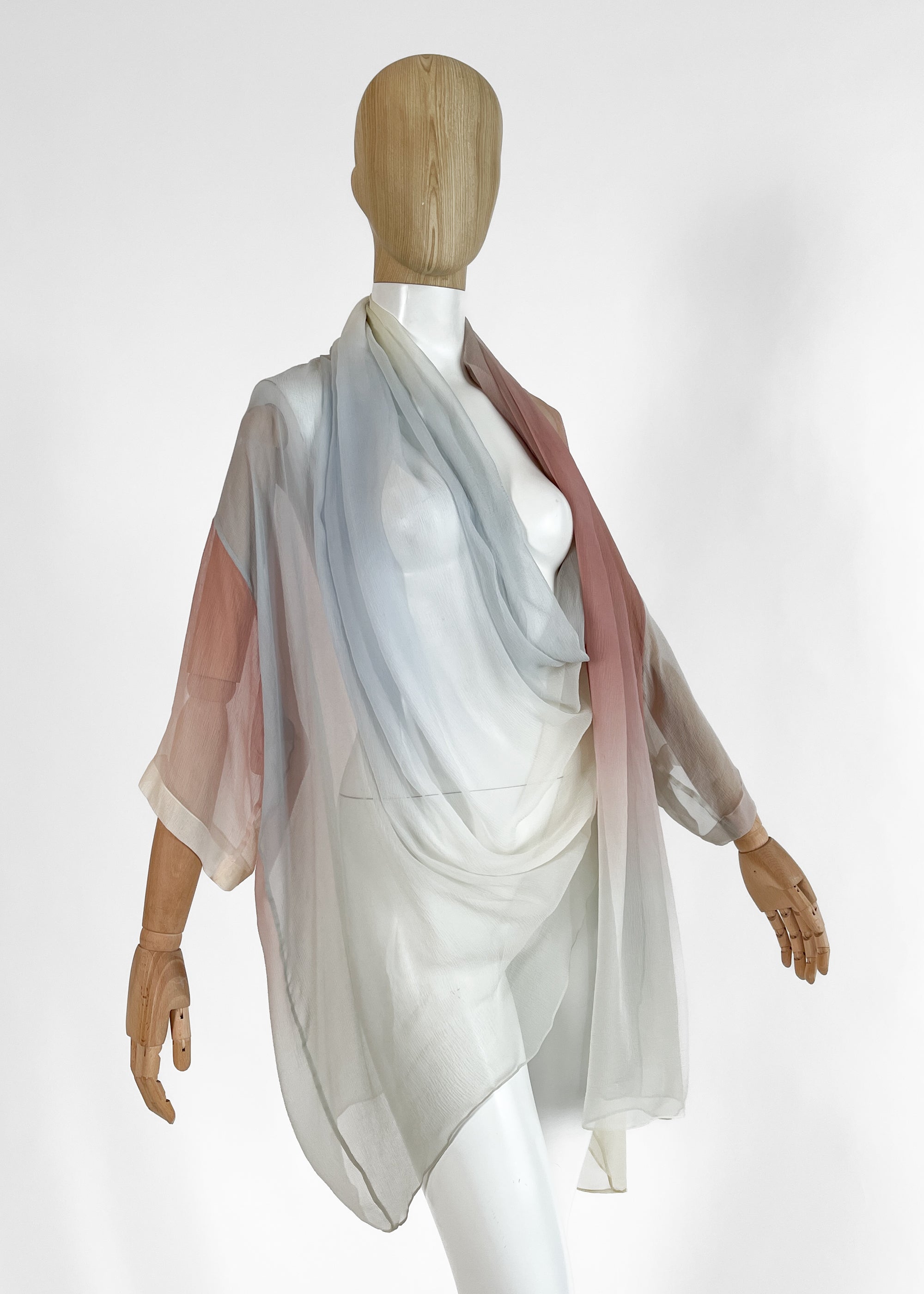 A 1989 Romeo Gigli sheer silk kimono-style wrap top draped on a mannequin. Features a soft pastel gradient transitioning from pale blue and grey to rose pink and ivory, with a draped cowl neckline and wide, fluid sleeves.  Photographed against a studio white brackground.
