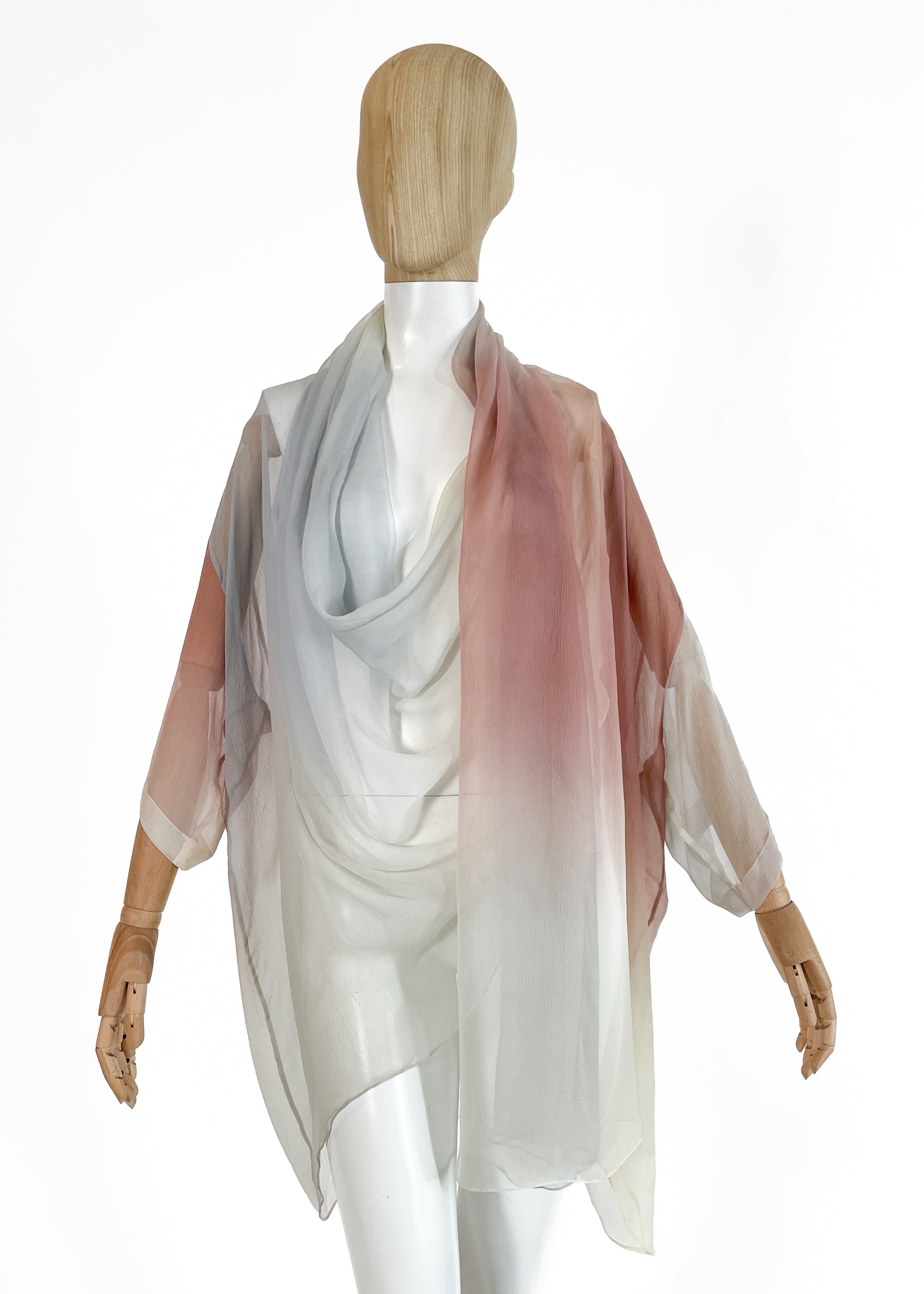 A 1989 Romeo Gigli sheer silk kimono-style wrap top draped on a mannequin. Features a soft pastel gradient transitioning from pale blue and grey to rose pink and ivory, with a draped cowl neckline and wide, fluid sleeves.  Photographed against a studio white background.